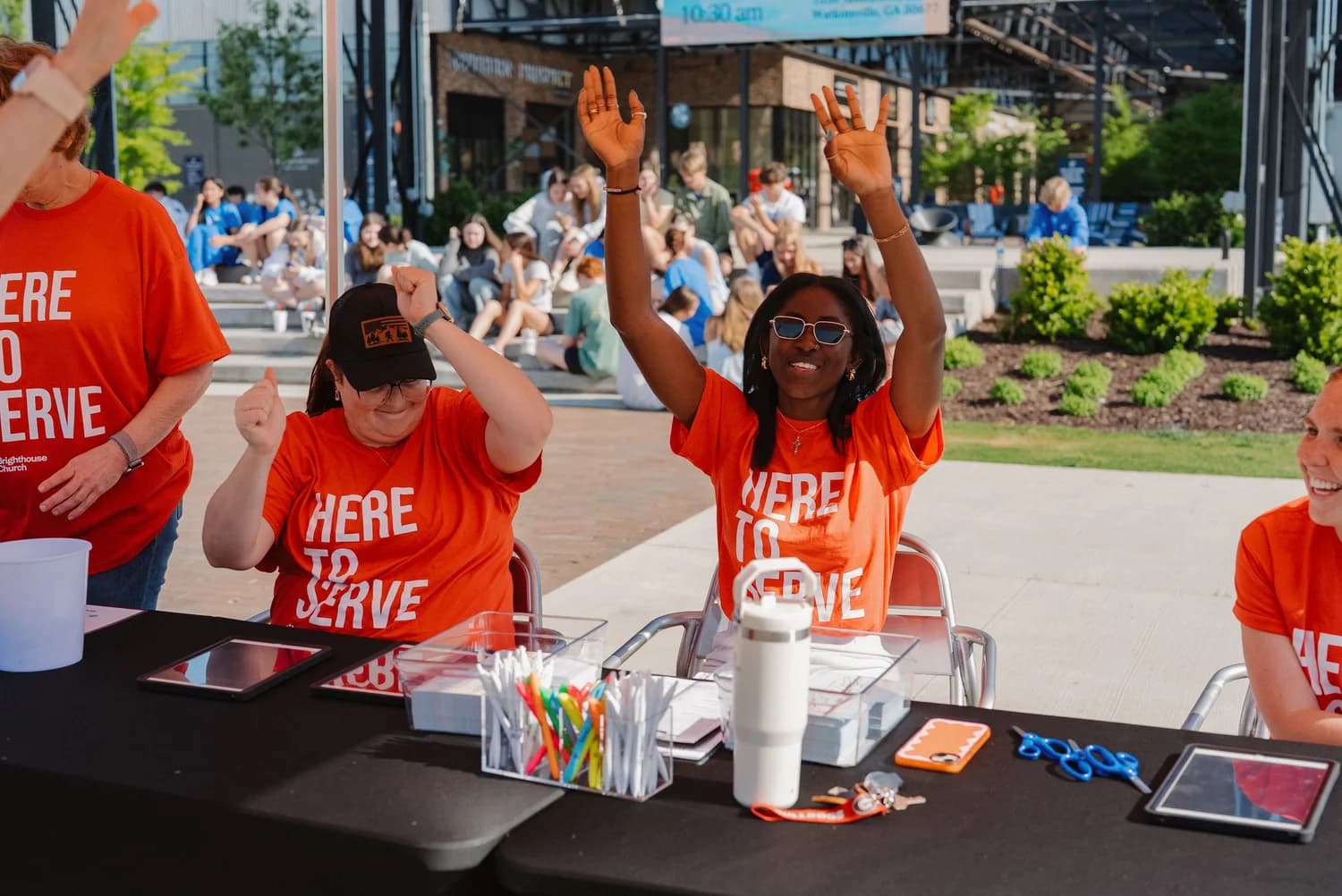 Brighthouse volunteers in orange Here to Serve shirts with arms raised at the welcome table